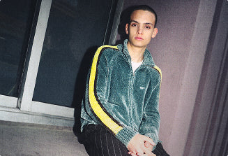 A person wearing a green velour tracksuit jacket with yellow stripes on the sleeves, sitting in a dimly lit indoor environment.
