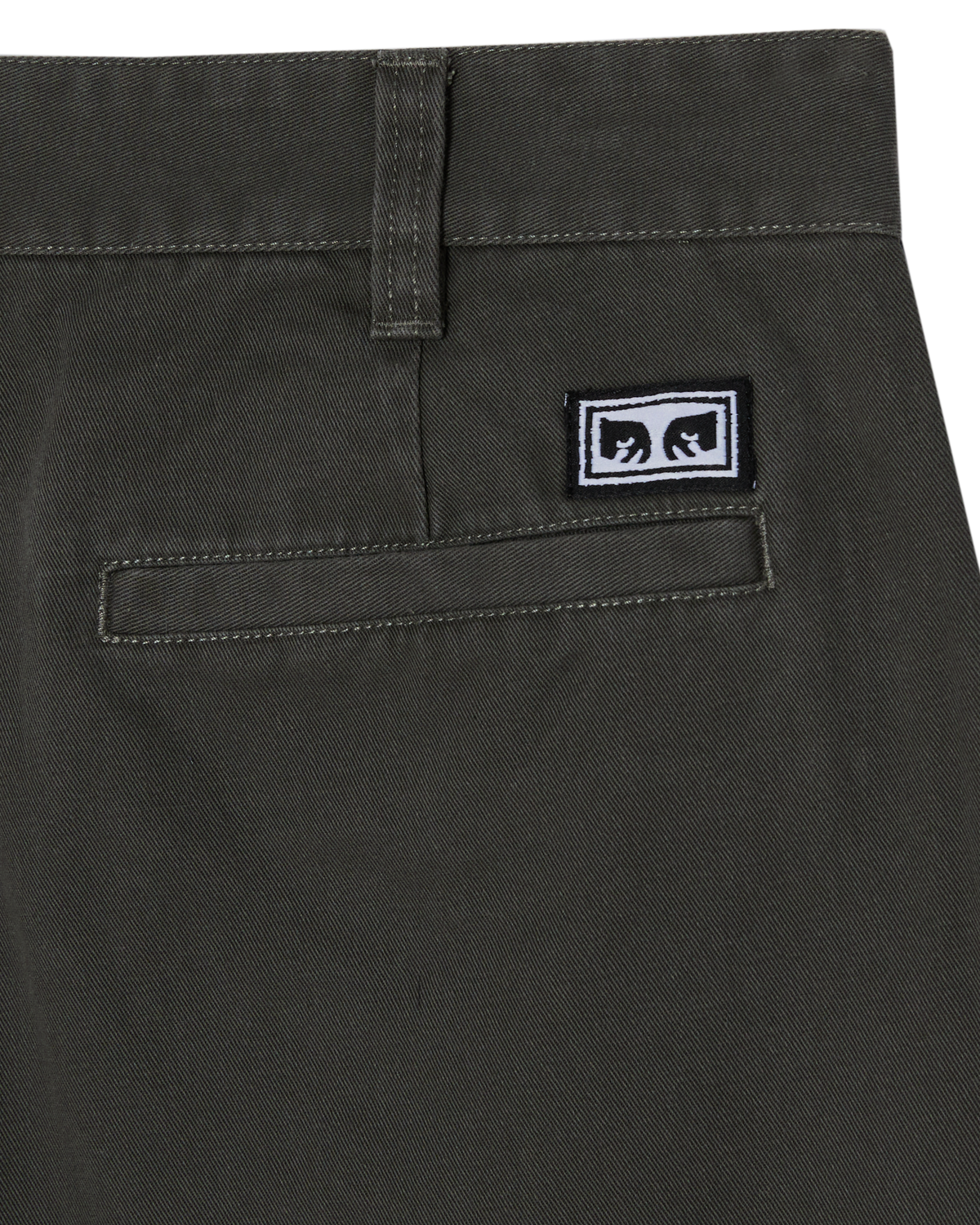 Close-up of the back waistband and pocket area of dark olive green pants, showing a small rectangular black-and-white logo patch on the pocket.
