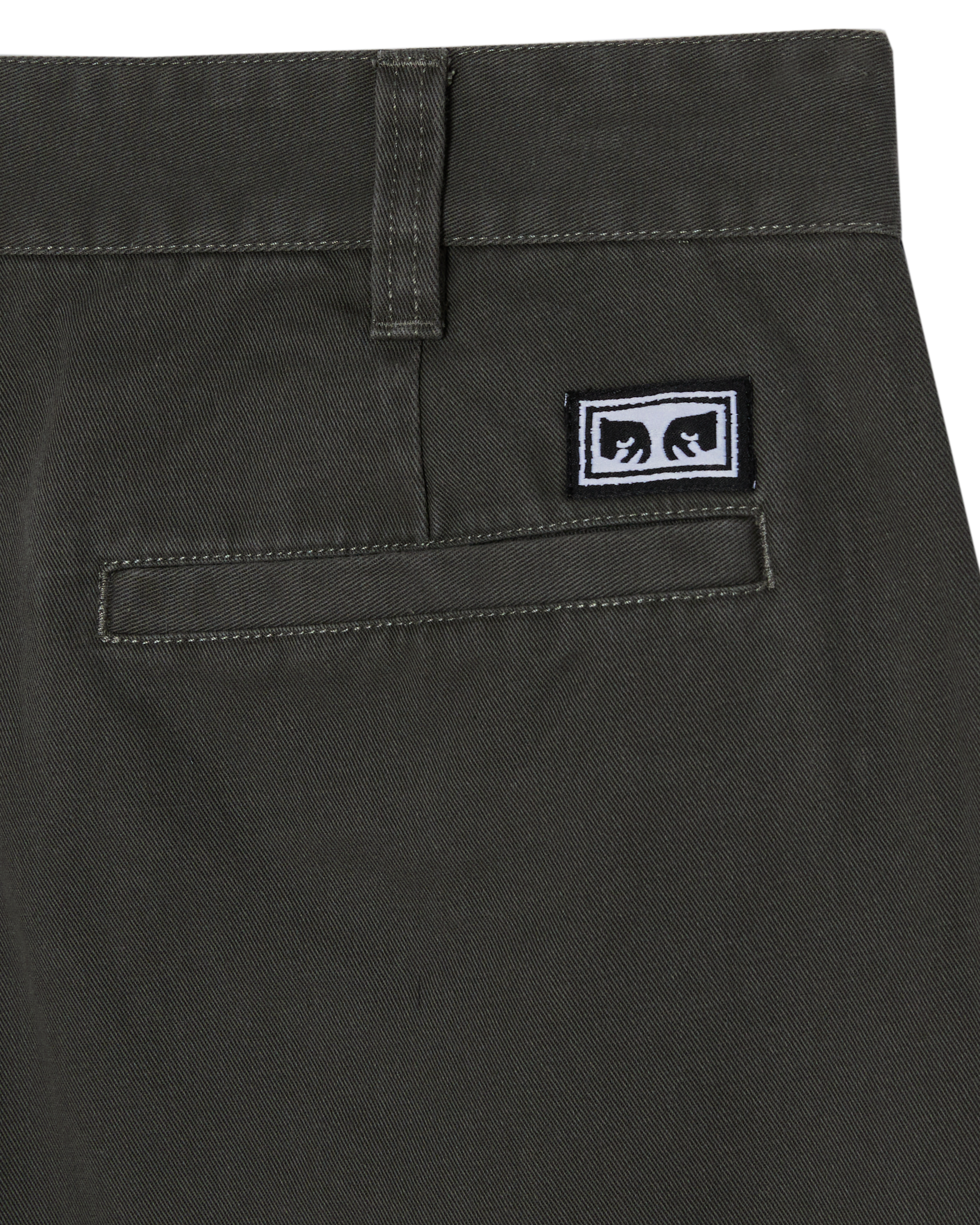 Close-up of the back waistband and pocket area of dark olive green pants, showing a small rectangular black-and-white logo patch on the pocket. (zoomed)