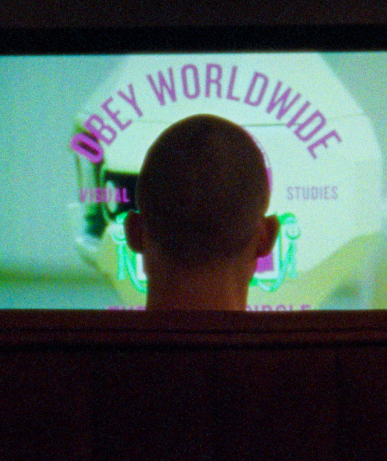 A person viewed from behind, seated in a dark room facing a large screen displaying the text 'OBEY WORLDWIDE' in purple over a pale green graphic.