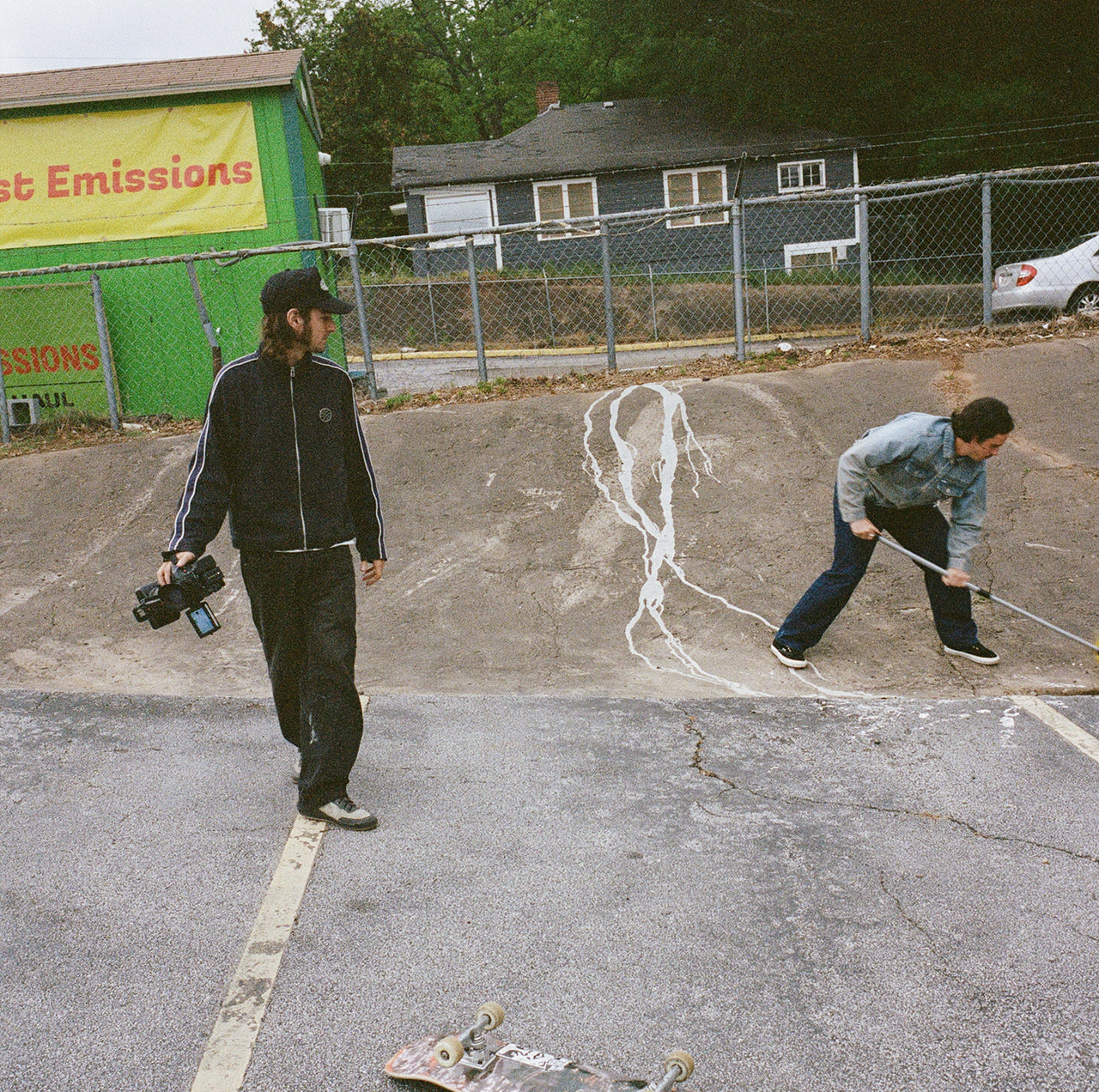 Two people in a parking lot with one sweeping the ground and the other holding a camera.