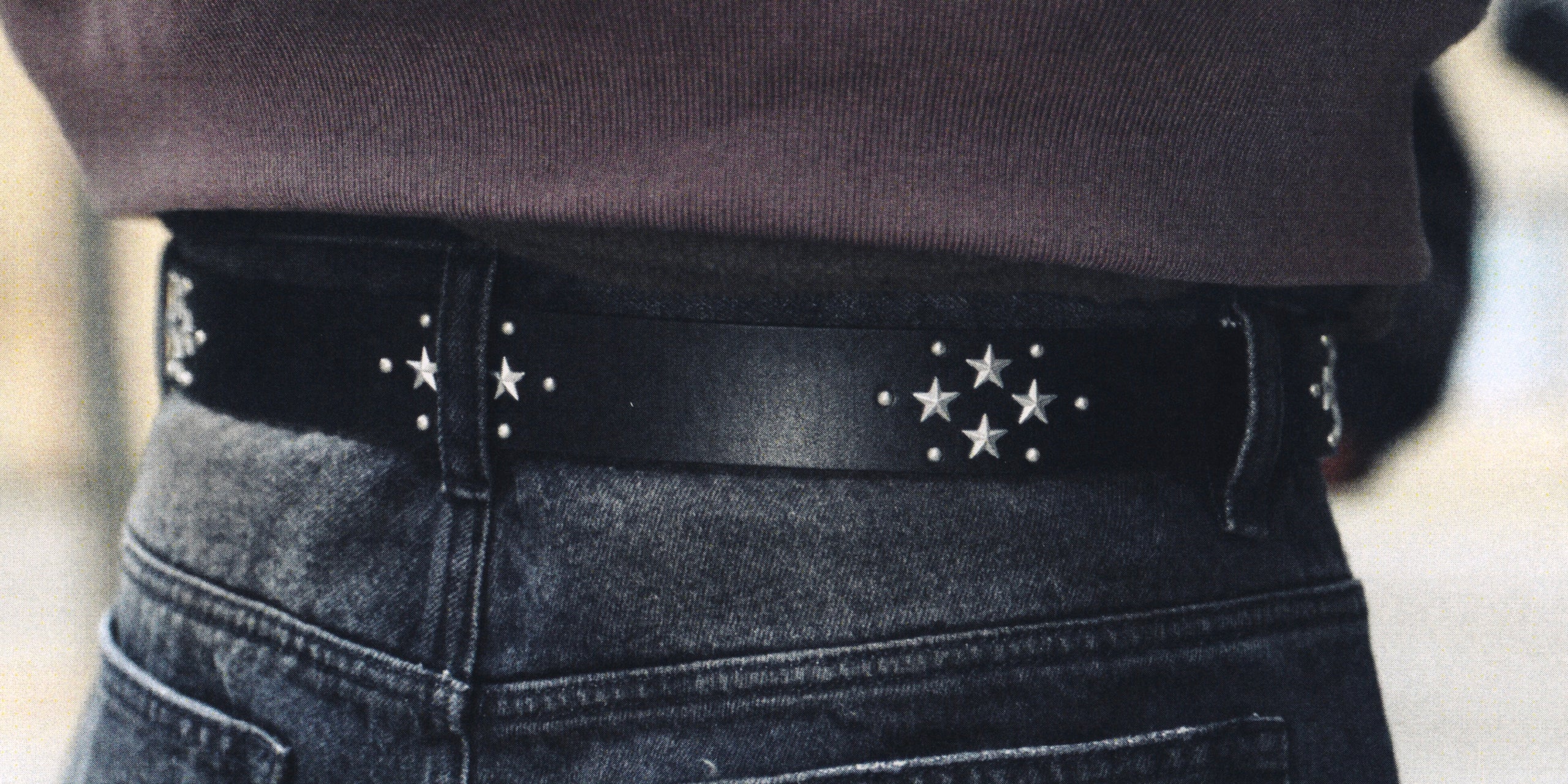 A close-up of a black leather belt with silver star and dot hardware, worn around the waist over dark gray jeans and a dark brown top.
