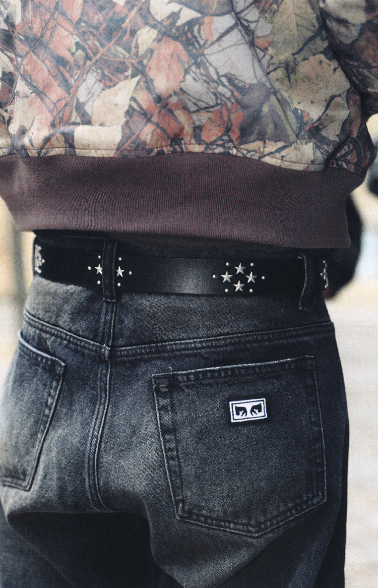Close-up of a person wearing dark gray washed denim jeans with a black studded belt and a brown ribbed hem, paired with a camouflage-patterned top.