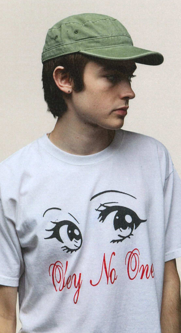 A young man wearing a light green baseball cap and a white graphic t-shirt with cartoon eyes and the red text 'Oleyn No One' against a plain beige background.