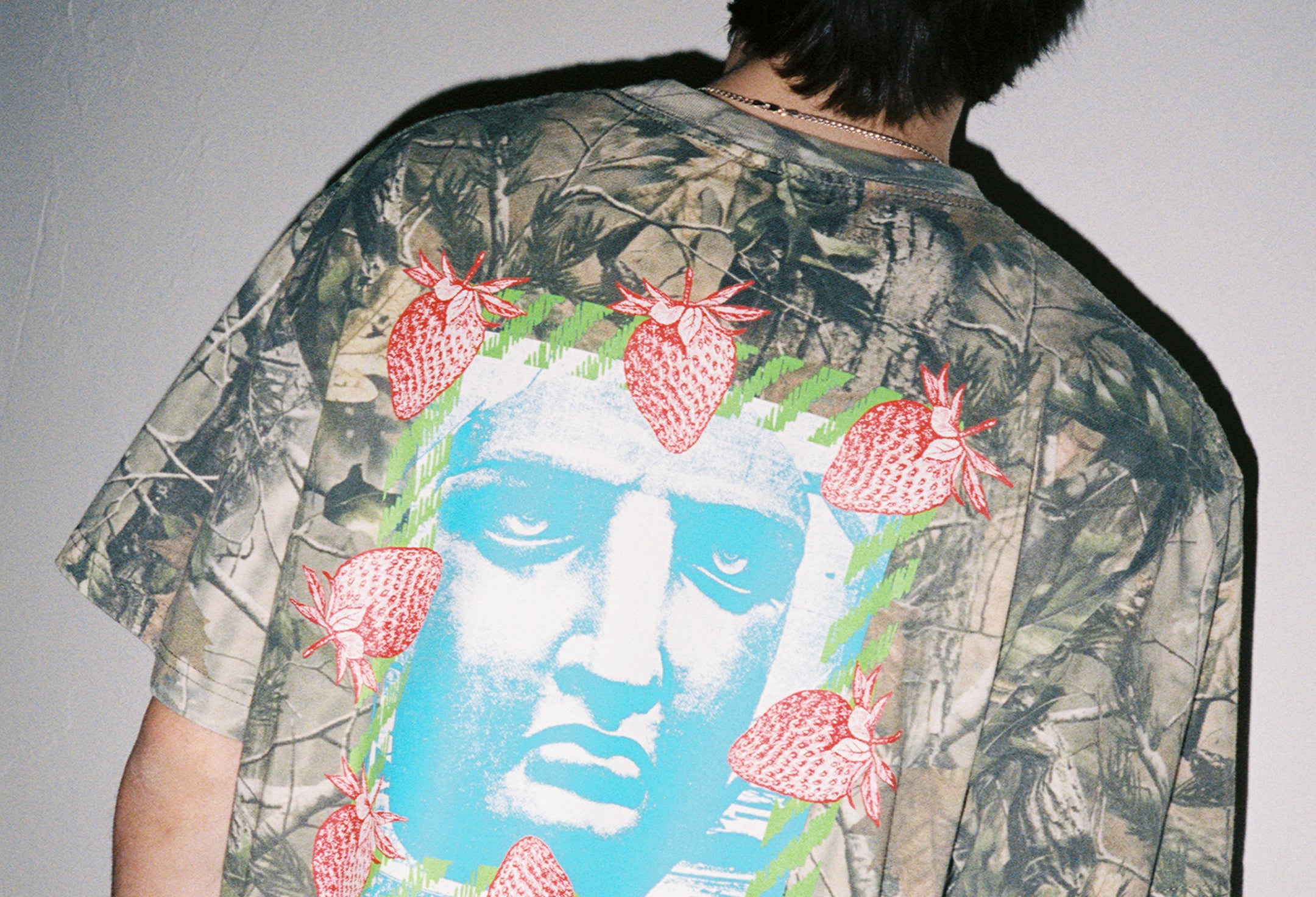 Person wearing a camouflage shirt with a face and strawberry design on the back.
