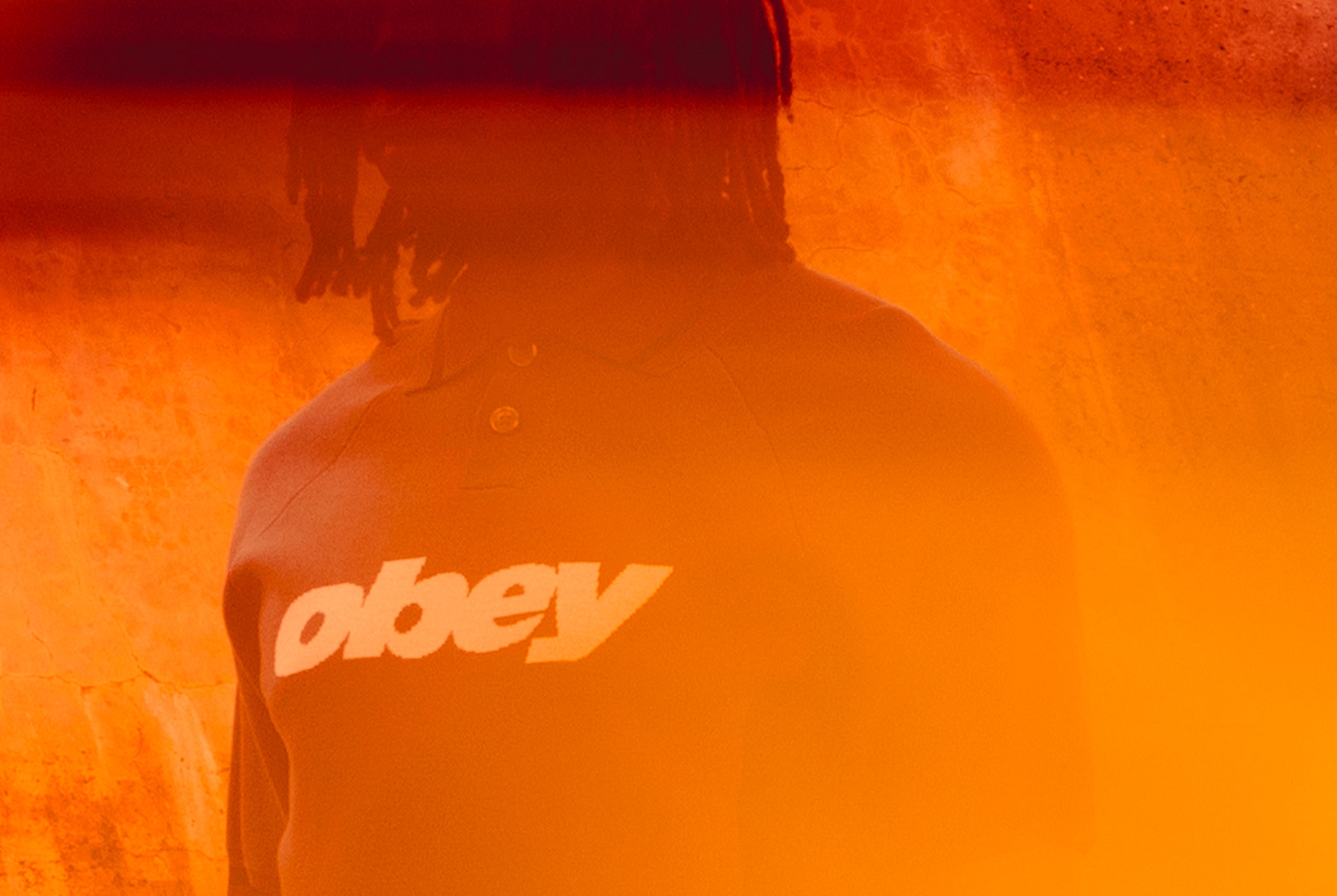 Person wearing a jacket with 'obey' logo in front of an orange background