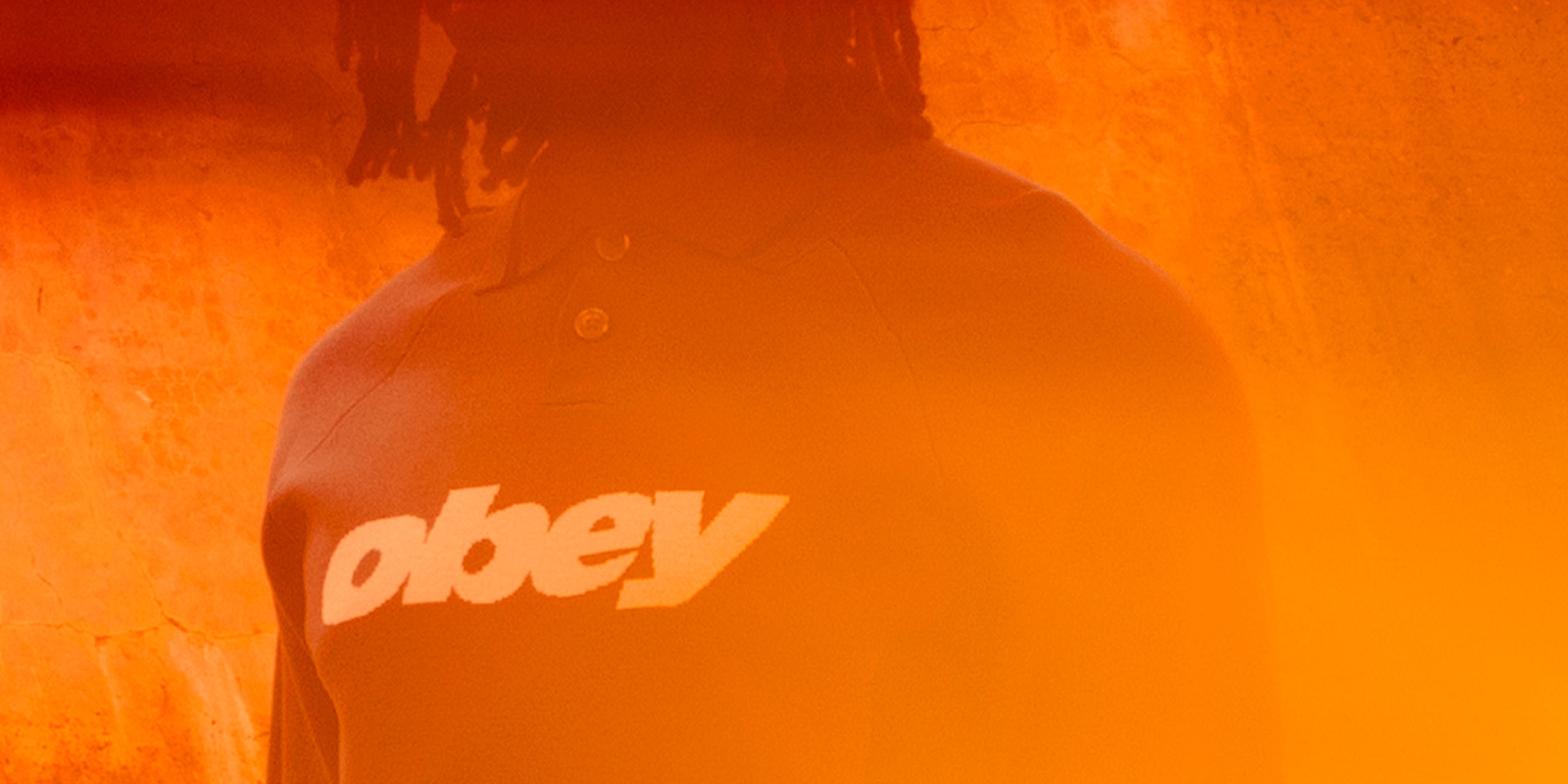 Person wearing a jacket with 'obey' logo in front of an orange background