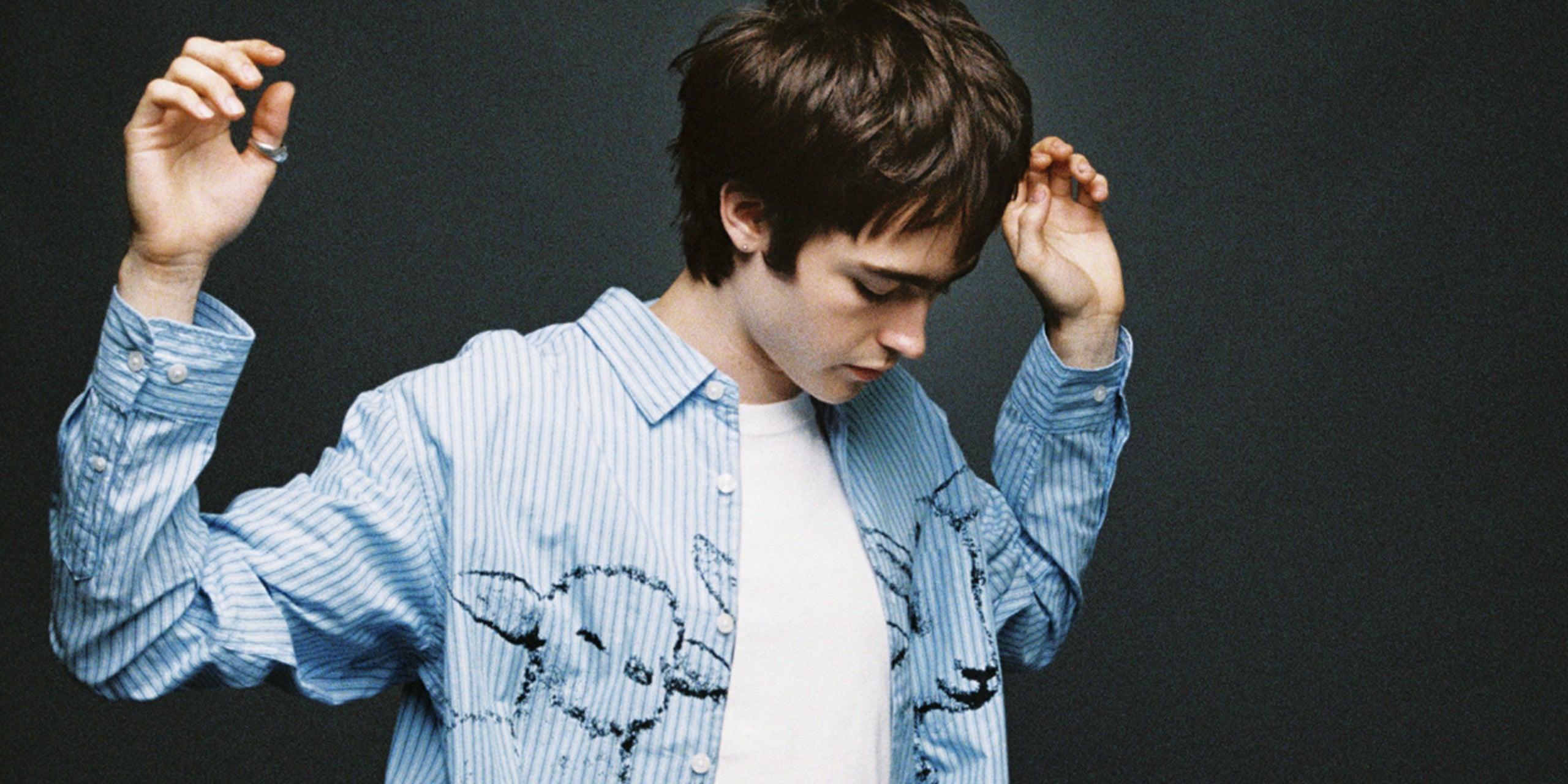 A young man with dark hair looking down, wearing a light blue and white vertically striped button-up shirt with a black graphic print on the front, layered over a white t-shirt, set against a dark gray background.
