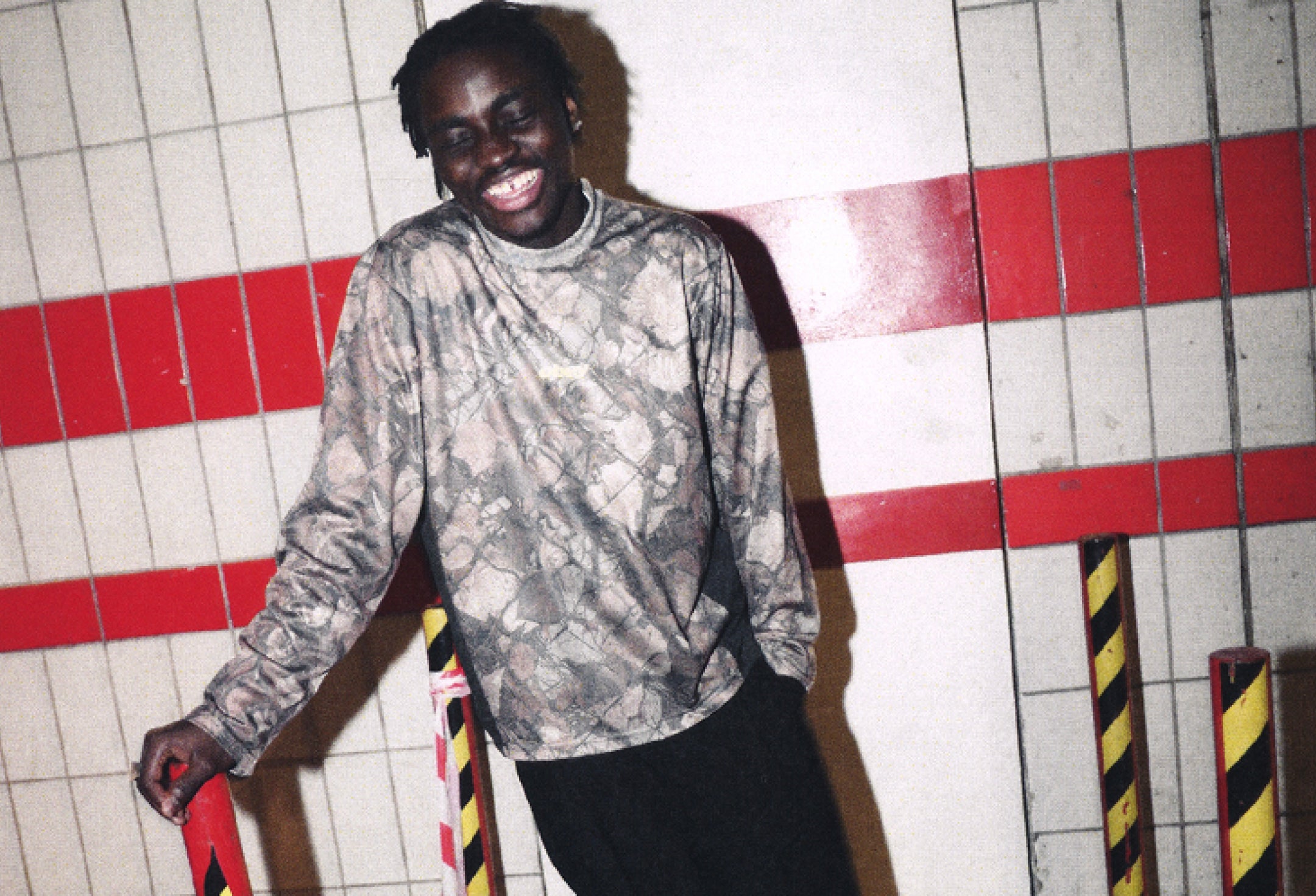 A man smiling and leaning against a tiled wall with red stripes, wearing a long-sleeve gray and black snakeskin-patterned shirt and black pants.
