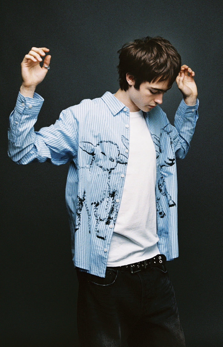 A man posing in a light blue and white vertically striped button-down shirt with black graphic prints, worn open over a white t-shirt and black jeans, set against a dark gray background.
