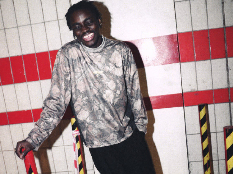 A man smiling and leaning against a tiled wall with red stripes, wearing a long-sleeve camouflage-patterned shirt and black pants.