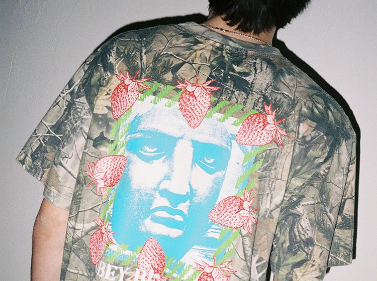 Person wearing a camouflage t-shirt with a graphic design of a face and strawberries.