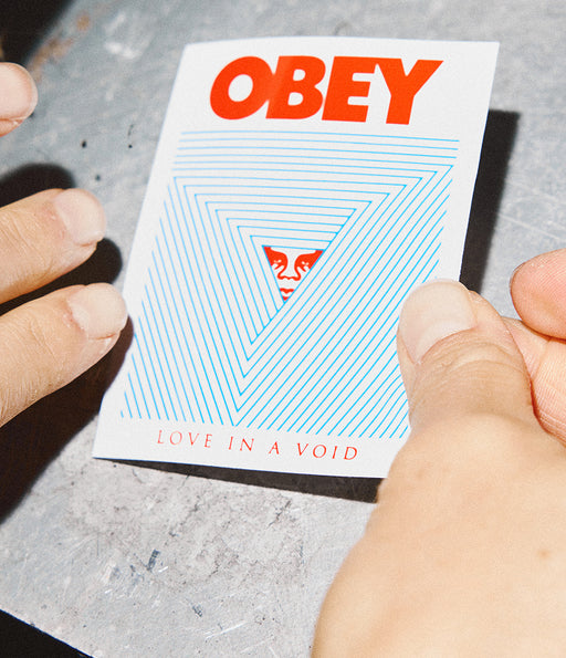 Card with 'OBEY' and 'Love in a Void' text held by hands on a gray surface