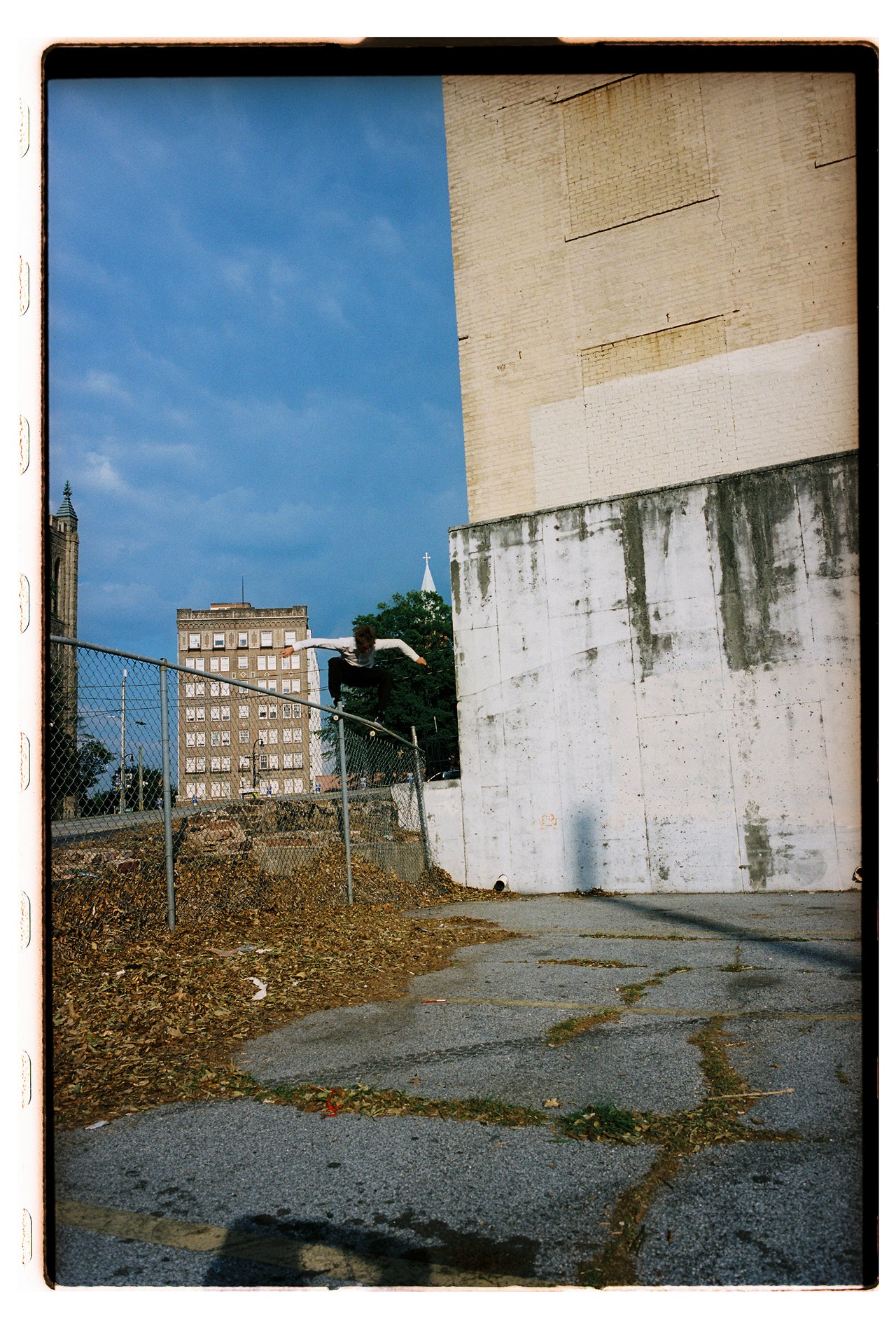 A square film-style photograph showing an urban outdoor scene with a large beige and white building wall, blue sky, chain-link fence, dry leaves, and cracked pavement. A person is visible jumping or standing near the fence in the middle distance.