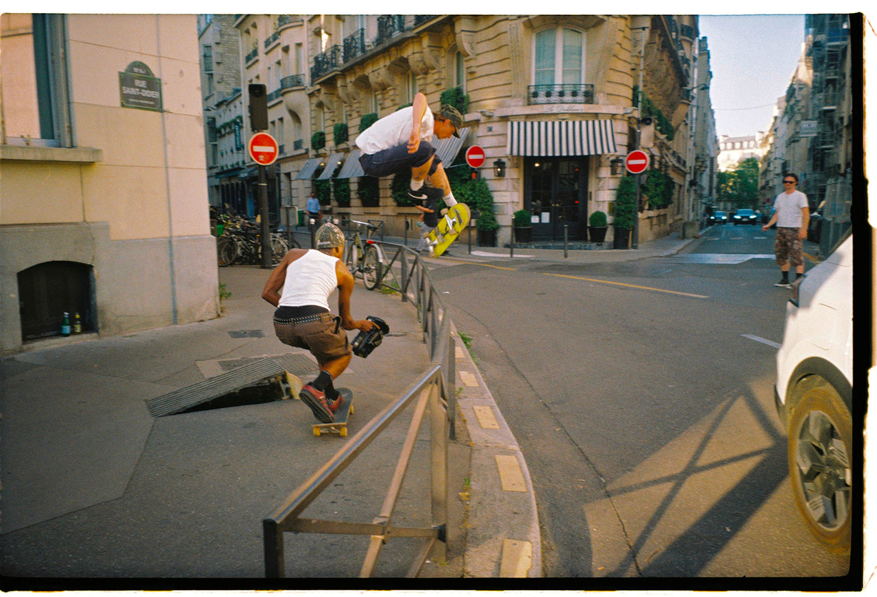A street scene with skateboarders in an urban city setting, featuring buildings, street signs, and a white car on the right. One skateboarder is airborne performing a trick while another grinds a metal rail in the foreground.