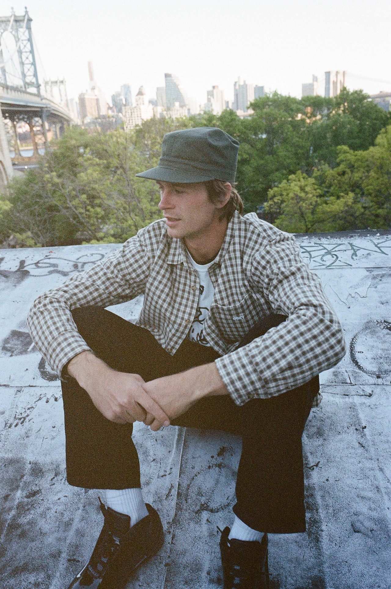 A man wearing a green cap, a beige and black checkered flannel shirt over a white t-shirt, dark pants, and black shoes, sitting on a concrete ledge with a city skyline and trees in the background.