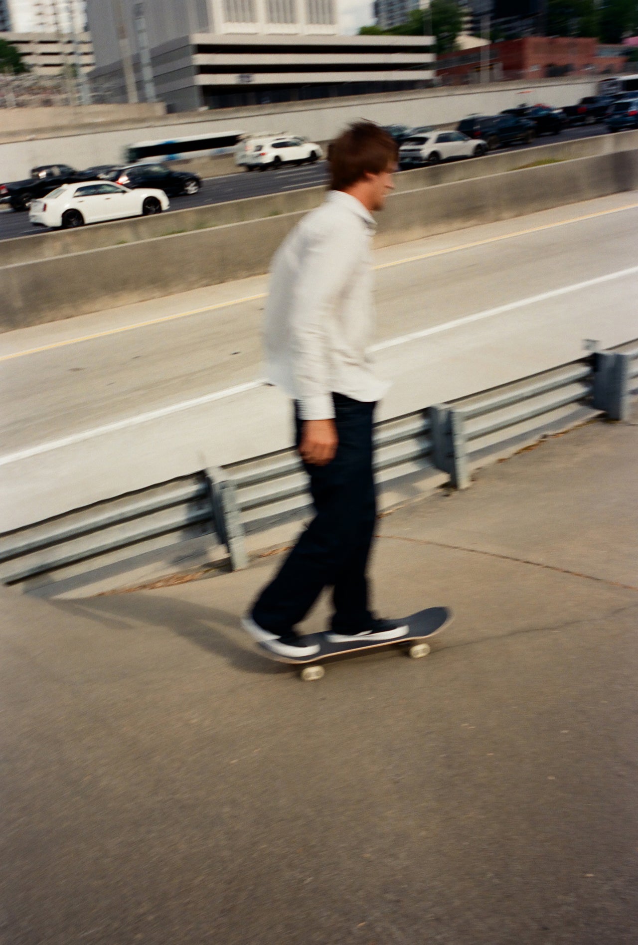 A person in a white long-sleeve shirt and dark pants rides a skateboard along a concrete path next to a highway with cars in the background.