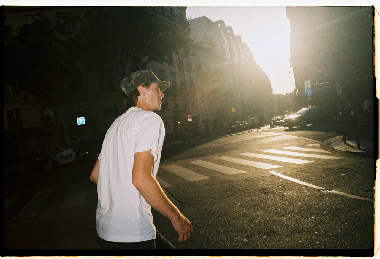 A man wearing a white t-shirt and a dark cap walks across a city street crosswalk during bright, low-angle sunlight with strong lens flare.