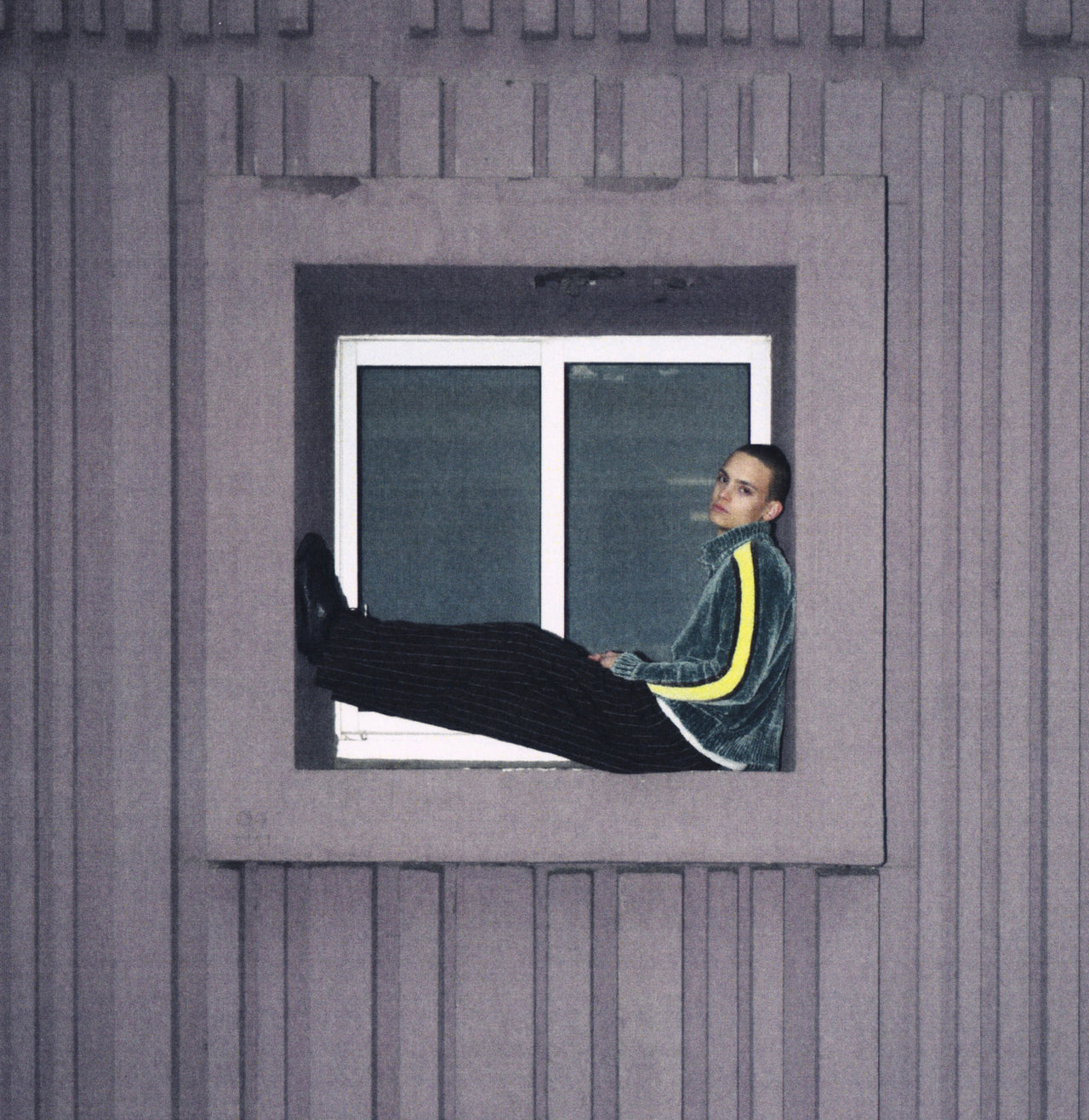 A person reclining in a window frame against a gray paneled exterior wall, wearing a dark green velvet tracksuit with a bright yellow stripe down the sleeve.