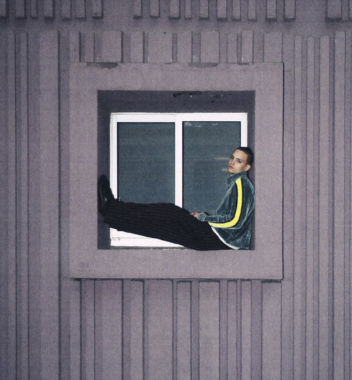A person wearing a dark green velvet tracksuit with a yellow stripe sits in a window of a gray ribbed building, with one leg stretched out.