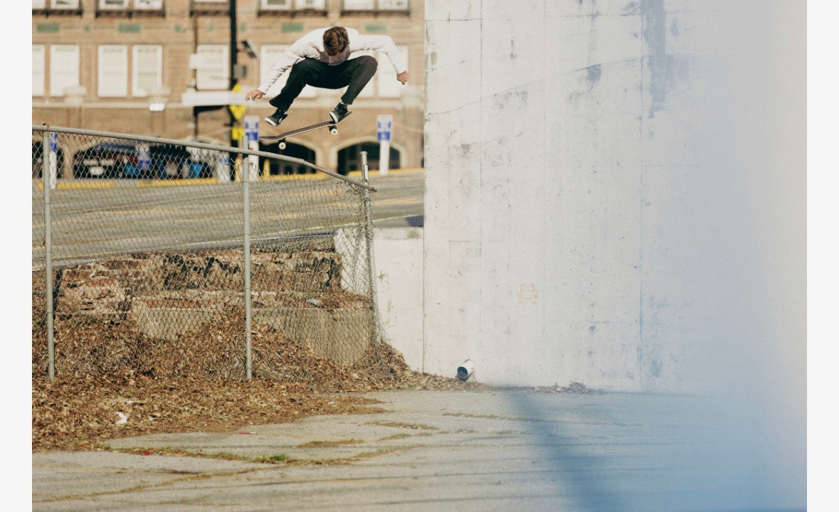 A skateboarder in mid-air performs a trick over a chain-link fence next to a large concrete wall, with an urban street and brick building visible in the background.