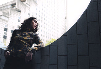 A person with curly dark hair wearing a camouflage-patterned long-sleeve shirt leans against a dark gray tiled wall in an outdoor urban setting with buildings in the background.